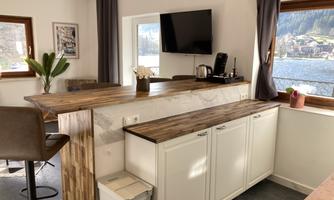 A modern kitchen with a wooden bar and white cabinets. In the background are windows overlooking a picturesque landscape.