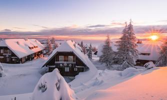 A winter landscape with snow-covered huts and trees. The sun rises on the horizon and bathes the scene in warm light.
