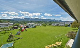 A large, green meadow with playground equipment for children and deck chairs. In the background, mountains and a clear, blue sky can be seen.