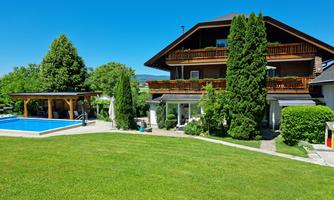 A beautiful house with several balconies and a large garden. In the background, there is a blue pool and lush trees.