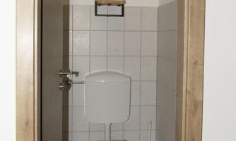 A small, clean bathroom with a toilet and a trash can. On the wall, there is a shelf with some decorative items.