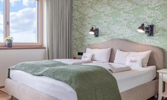 A cozy bedroom with a large bed and a green bedspread. The walls are decorated with a modern, botanical pattern.