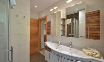 A modern bathroom with a large double marble sink and wooden shelves. The shower is separated by a glass wall and the room is brightly lit.