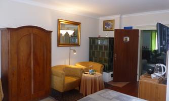 Room with wooden wardrobe, seating area with two armchairs and round table, tiled stove, mirror, floor lamp, tea station and view into the central hall from which all rooms are accessed.