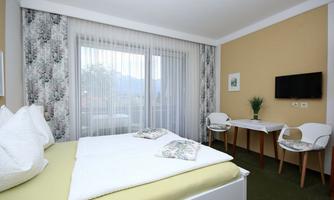 A modern bedroom with a large bed and inviting decor. The room has large windows overlooking nature and a small seating area.