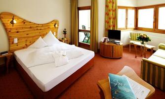 A cozy hotel room with a large bed and wooden elements. There is a seating area, a television, and plenty of daylight through the windows.
