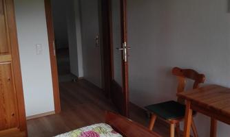 A simple bedroom with a wooden bed, a table, and a chair. The walls are light, and there is a door that leads to another room.