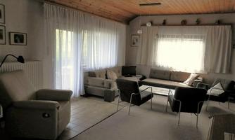 A cozy living room with a wooden ceiling and large windows. The furniture is modern and inviting, perfect for relaxing.