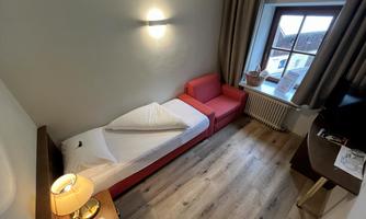 A simple room with a single bed and a red couch. The room has a large window with a view outside.