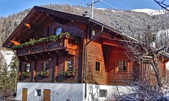 A beautiful, traditional wooden house in the mountains, surrounded by snow. The balconies are decorated with plants, and the sky is clear and blue.