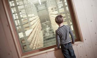 Ein Junge steht vor einem Fenster und schaut auf Regale mit Käse. Der Raum hat eine warme, einladende Atmosphäre.