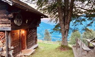 A picturesque wooden cabin overlooking the valley and the mountains. In the foreground, there is a wooden bench and trees that create a peaceful atmosphere.