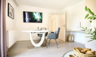 A bright and modern living room with a table, chairs, and a TV on the wall. On a side table, there are cookies in a basket and fresh flowers.
