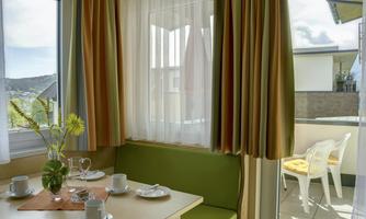A cozy dining area with a table, dishes, and a flower. Bright curtains and a view of the balcony create an inviting atmosphere.