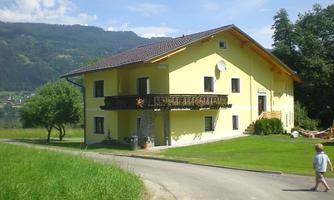 A yellow house with a balcony and a green lawn area. In the background, mountains and trees can be seen.