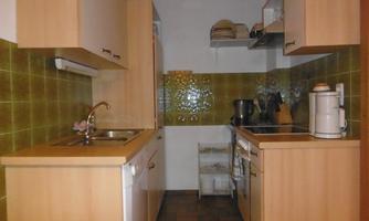 A small kitchen with wooden cabinets and green tiles. Cooking appliances and dishes are visible on the countertop.