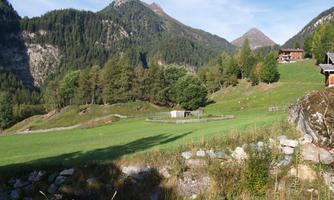 A picturesque landscape with gentle hills and tall mountains in the background. In the foreground, there is a green meadow with trees and a small wooden building.
