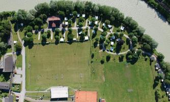 An aerial view of a campsite by the river with many pitches and a large green lawn. Surrounded by trees and paths, the environment invites relaxation.