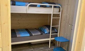 A simple bedroom with a bunk bed and a small table. The walls are made of wood, and the floor has grey tiles.