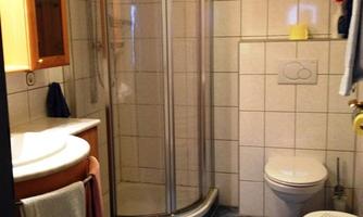 A small bathroom with a shower and a toilet. There is a sink area with towels and a small stool.