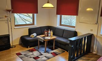 A cozy living area with a black sofa and a small table. Daylight streams in through two windows, and the room is decorated with red curtains.