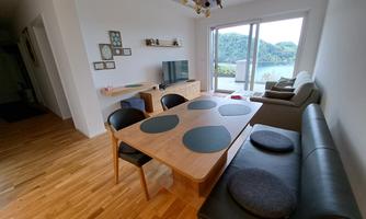 A modernly furnished living area with a wooden table and a cozy sofa. Large windows offer a view of the green landscape and the water.