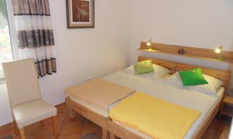 A cozy room with a double bed and two colorful pillows. A chair is next to the bed and the window lets in daylight.