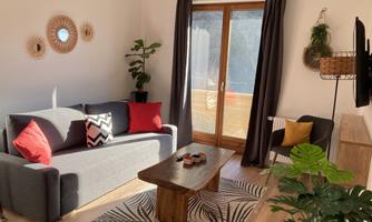 A modern living room with a gray couch and red cushions. The room is bright and inviting, with plants and a large window.