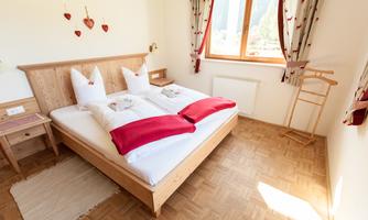 A cozy bedroom with a double bed and two pillows in white and red. Large windows let in plenty of light, and the walls are decorated with pretty curtains.
