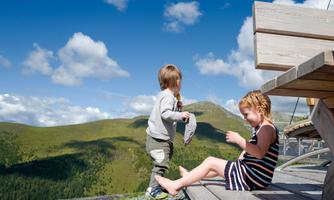 Two children are playing outside on a terrace with a view of green mountains. The sky is clear and blue with a few clouds.