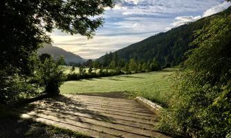 A picturesque landscape with gentle hills and a clear sky. A wooden path leads through the green meadow to nature.