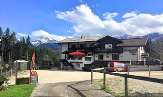 A cozy inn surrounded by trees and mountains. The sky is blue and there is an inviting terrace with a red sunshade.