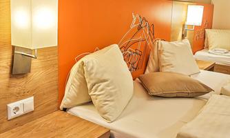 A modern bedroom with two single beds and a warm orange wall. The decor is simple and cozy with pillows and nightstands.