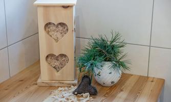 A wooden box with heart cutouts is sitting on a table, surrounded by pine needles in a decorative pot. Next to the box are wood shavings and a gray felt heart.