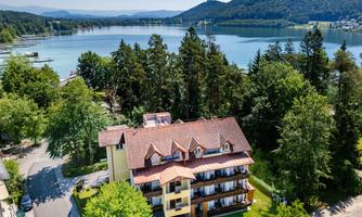 A hotel building surrounded by trees and green meadows, overlooking a clear lake. Gentle hills can be seen in the background.