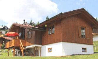 A traditional wooden house in a green landscape. It has a balcony with a red sunshade and is surrounded by other houses.