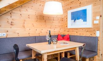 A cozy dining area with wooden walls and a large table. On the table are wine and glasses, surrounded by comfortable benches and chairs.