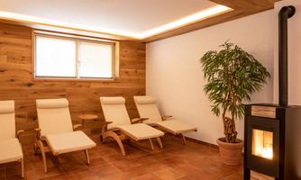 A tranquil wellness area with cozy loungers and a wood stove. The room features wooden walls and a plant arrangement that creates a relaxing atmosphere.