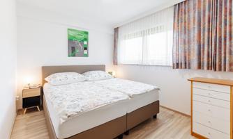 A bright bedroom with a double bed and a dresser. The windows are decorated with colorful curtains and a picture frame hangs on the wall.