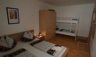 A bright, cozy bedroom with a double bed and a bunk bed. The furnishings are simple and modern, with wooden flooring and minimalist decor.