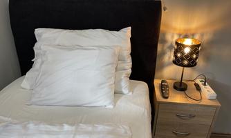 A cozy bedroom with a bed that has two pillows. Next to the bed is a nightstand with a lamp and a remote control.