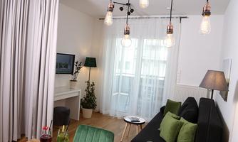A modern living room with a cozy couch and green cushions. Large windows let in plenty of light and the room is decorated with stylish lamps and plants.