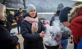 Eine Gruppe von Menschen steht im Freien und genießt eine winterliche Atmosphäre. Eine Frau bereitet Speisen in einem Topf über einem Feuer, während andere mit Laternen in der Hand um sie herumstehen.