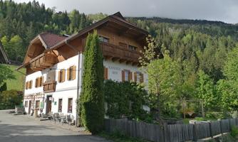 A charming two-story building with a wooden porch and shutters. Surrounded by green trees and mountains, it radiates coziness.