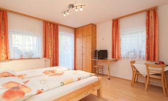 A cozy bedroom with a double bed, wooden furniture, and large windows. The room is bright and friendly, with attractive orange curtains.