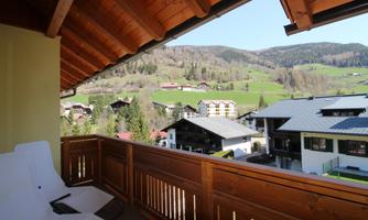 A balcony with wooden railing and cozy loungers. In the background, a green landscape stretches out with mountains and several houses.