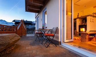 A cozy terrace with chairs and a table on one side and a warm living area on the other. In the background, snow-covered mountains and a clear sky are visible.