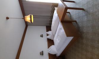 A simple room with two single beds and a small table. There is a large window front with curtains and a ceiling lamp.