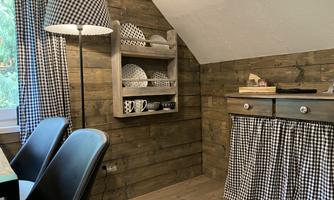A cozy room with wooden walls and a window. There is a table, chairs, and a shelf with dishes as well as a lamp in the same pattern.