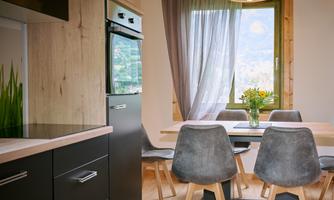 A modern kitchen with a dining table and comfortable chairs. Daylight falls through the window and there is a flower arrangement on the table.